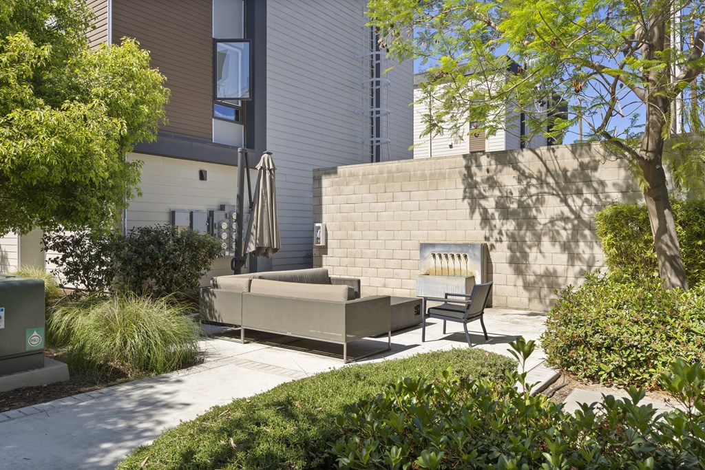 A patio with a couch and chairs is surrounded by green bushes.
